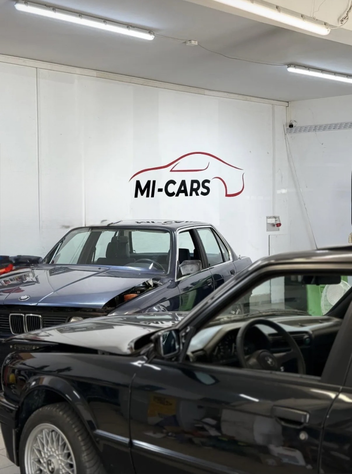 Hala serwisowa MI-CARS Auto Serwis z podświetlonym logo MI-CARS na ścianie i dwoma klasycznymi BMW E30 przygotowanymi do obsługi na stanowisku warsztatowym